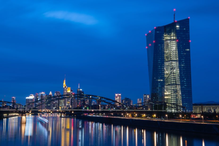 Ein vorbeifahrendes Schiff zieht kurz nach Sonnenuntergang seine Lichtspuren über den Main, während im Hintergrund die Lichter der Frankfurter Bankenskyline und der Europäischen Zentralbank (EZB) leuchten.