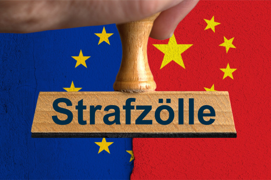 Ein symbolischer Holzstempel mit der Aufschrift "Strafzoelle", gehalten von einer Hand im Anschnitt vor der Flagge der Europaeischen Union und der Nationalflagge der Volksrepublik China auf einer Wand mit einem vertikalen Absatz bzw. Riss in der Mitte und zwei unterschiedlichen Putzarten.