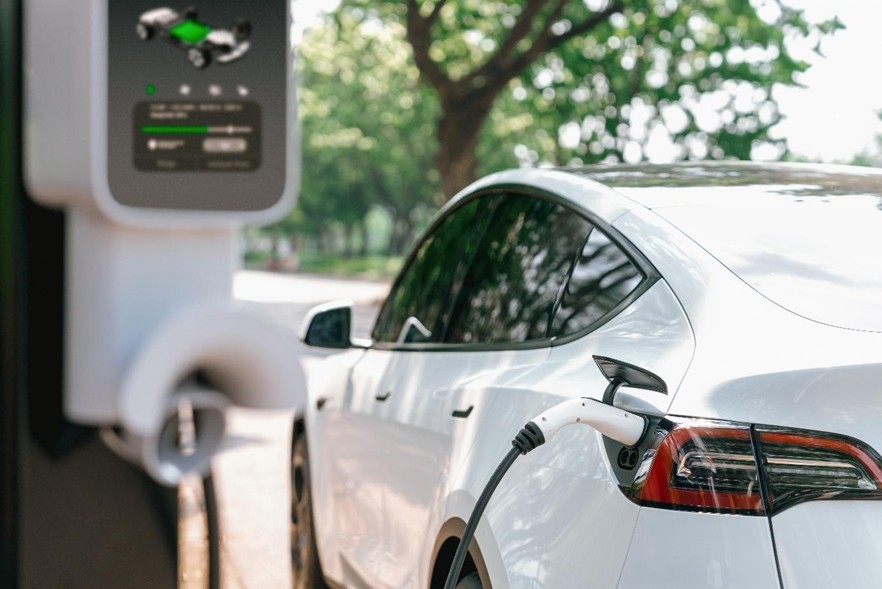 Ein weißes Elektroauto befindet sich neben einer Ladestation. Im Hintergrund sind Bäume zu sehen.